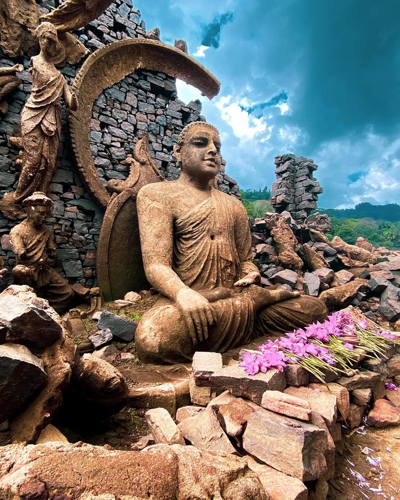 Stone statue of a seated Buddha among ancient brick ruins, with pink flowers placed as offerings